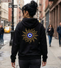 A woman viewed from behind wearing a black hoodie with a large gold sunburst and blue-and-white eye graphic on the back, walking on a city street.