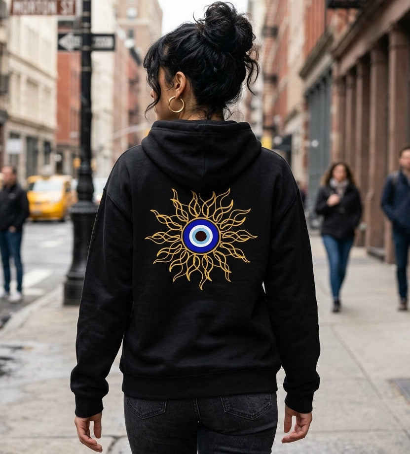 A woman viewed from behind wearing a black hoodie with a large gold sunburst and blue-and-white eye graphic on the back, walking on a city street.