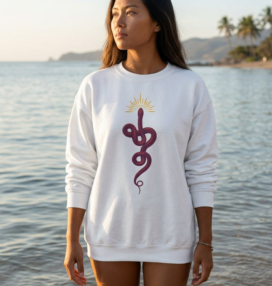 A woman wearing a white crewneck sweatshirt with a dark purple caduceus graphic and a yellow sunburst, standing by the ocean with a beach and palm trees in the background.